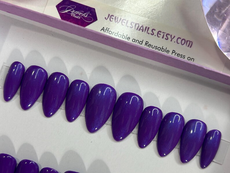 Puede incluir: Un conjunto de diez u&ntilde;as postizas de color morado con un acabado brillante. Las u&ntilde;as est&aacute;n dispuestas en una fila sobre una superficie blanca. JEWELSNAILS.ETSY.COM est&aacute; impreso en una caja blanca en el fondo. Presiona sobre asequible y reutilizable.