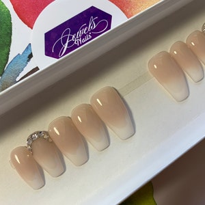 May include: A set of ten nude-colored, long, coffin-shaped press-on nails with a white ombre effect. The nails have a silver rhinestone accent on the top of the nail.