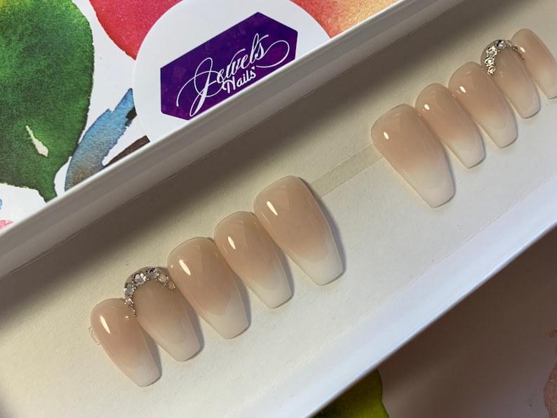 May include: A set of ten nude-colored, long, coffin-shaped press-on nails with a white ombre effect. The nails have a silver rhinestone accent on the top of the nail.
