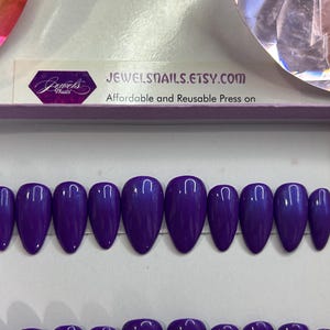 Puede incluir: Un conjunto de diez u&ntilde;as postizas de color morado con un acabado brillante. Las u&ntilde;as tienen forma de stiletto y est&aacute;n dispuestas en fila sobre una superficie blanca. JEWELSNAILS.ETSY.COM Affordable and Reusable Press on
