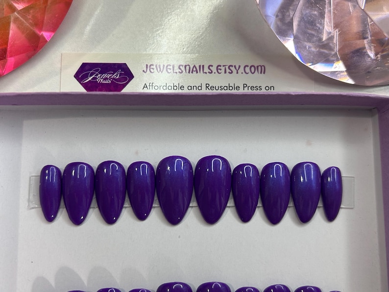 Puede incluir: Un conjunto de diez u&ntilde;as postizas de color morado con un acabado brillante. Las u&ntilde;as tienen forma de stiletto y est&aacute;n dispuestas en fila sobre una superficie blanca. JEWELSNAILS.ETSY.COM Affordable and Reusable Press on
