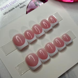 Könnte beinhalten: Zwei Sets von aufklebbaren Nägeln mit einem klassischen French-Maniküre-Design. Jeder Nagel hat eine glänzende, hellrosa ovale Form mit einer weißen Spitze. Die Nägel werden auf einer weißen Karte mit dem Text "JEWELSNails Affordable and Reusable Nails" präsentiert.