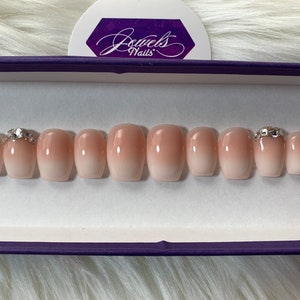 May include: A set of ten nude ombre press-on nails with a clear base and a light pink gradient. Two of the nails have a small, clear rhinestone accent.