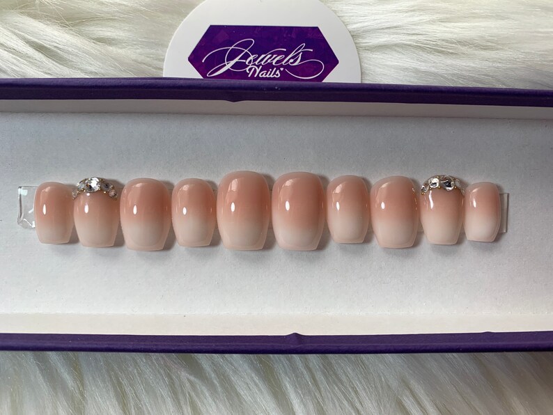 May include: A set of ten nude ombre press-on nails with a clear base and a light pink gradient. Two of the nails have a small, clear rhinestone accent.