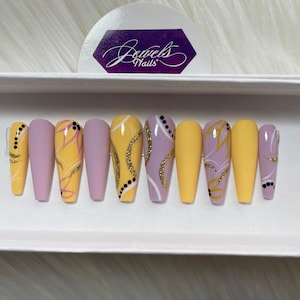 May include: Set of ten false nails with a lavender and yellow base and abstract gold and black designs. The nails are displayed in a white box with the text "Jewels Nails" on a sticker.