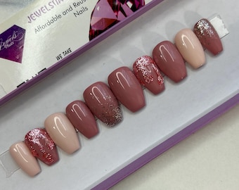 May include: Set of ten false nails with a pink and nude base and glitter accents. The nails are displayed in a purple box with the text "Jewelsta Affordable and Reusable Nails" and "We Take".