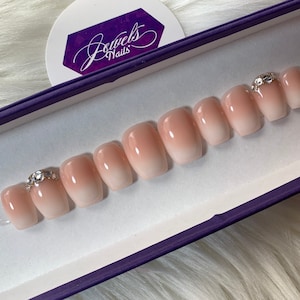 May include: A set of ten nude-colored, ombre press-on nails with a clear base and a pink gradient. The nails are arranged in a row in a purple box with the text "Jewels Nails" on the lid. Each nail has a small, clear rhinestone on top.