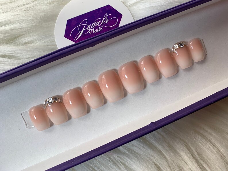 May include: A set of ten nude-colored, ombre press-on nails with a clear base and a pink gradient. The nails are arranged in a row in a purple box with the text "Jewels Nails" on the lid. Each nail has a small, clear rhinestone on top.