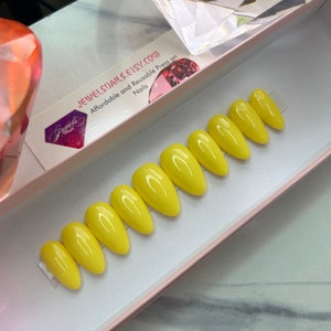 May include: A set of ten yellow almond-shaped press-on nails in a pink box. The box has the text "JewelSnails Etsy.com Affordable and Reusable Press on Nails" printed on it.