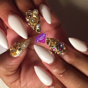 May include: White stiletto nails with gold and crystal embellishments. The nails are long and pointed, and the embellishments are clustered around the base of each nail.