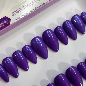 Puede incluir: Un conjunto de 10 u&ntilde;as postizas de stiletto de color morado con un acabado brillante. Las u&ntilde;as est&aacute;n dispuestas en dos filas sobre una superficie blanca. El texto "JEWELSNAILS.ETC" es visible en la esquina superior izquierda de la imagen. El texto "Affordable and Reusable Press-On Nails" tambi&eacute;n es visible en la esquina superior izquierda de la imagen.
