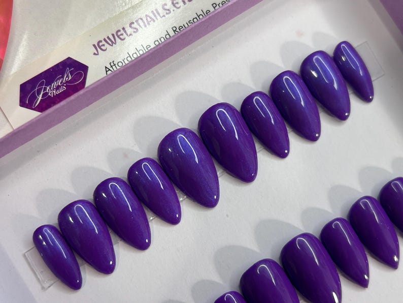 Puede incluir: Un conjunto de 10 u&ntilde;as postizas de stiletto de color morado con un acabado brillante. Las u&ntilde;as est&aacute;n dispuestas en dos filas sobre una superficie blanca. El texto "JEWELSNAILS.ETC" es visible en la esquina superior izquierda de la imagen. El texto "Affordable and Reusable Press-On Nails" tambi&eacute;n es visible en la esquina superior izquierda de la imagen.