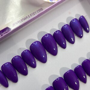 Puede incluir: Un conjunto de diez u&ntilde;as postizas de stiletto de color morado con un acabado brillante. Las u&ntilde;as est&aacute;n dispuestas en dos filas sobre una superficie blanca. El texto "JEWELSTAILS.ETSY.COM" es visible en el fondo.