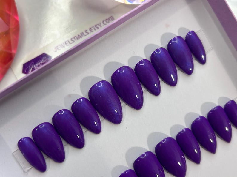 Puede incluir: Un conjunto de diez u&ntilde;as postizas de stiletto de color morado con un acabado brillante. Las u&ntilde;as est&aacute;n dispuestas en dos filas sobre una superficie blanca. El texto "JEWELSTAILS.ETSY.COM" es visible en el fondo.