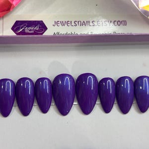 Puede incluir: Diez u&ntilde;as postizas de almendra de color morado con un acabado brillante. Las u&ntilde;as est&aacute;n dispuestas en una fila sobre una superficie blanca.