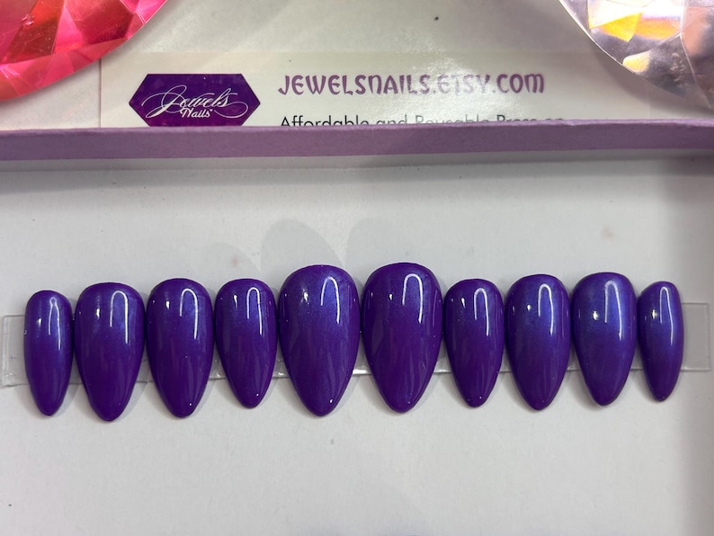 Puede incluir: Diez u&ntilde;as postizas de almendra de color morado con un acabado brillante. Las u&ntilde;as est&aacute;n dispuestas en una fila sobre una superficie blanca.
