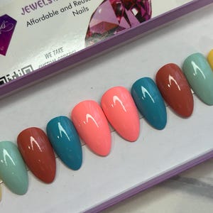 May include: A set of ten press-on nails in various colors, including yellow, light blue, teal, brown, pink, and red. The nails are arranged in a row on a white surface.