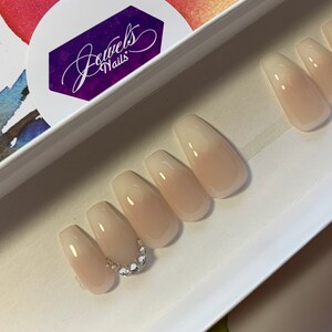 May include: A set of ten nude-colored coffin-shaped press-on nails with a gradient effect. The nails have a clear base with a light pink color that fades to a darker pink at the tip. The nails are decorated with a row of small rhinestones on the accent nail.