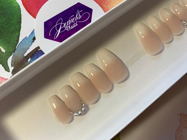 May include: A set of ten nude-colored coffin-shaped press-on nails with a gradient effect. The nails have a clear base with a light pink color that fades to a darker pink at the tip. The nails are decorated with a row of small rhinestones on the accent nail.