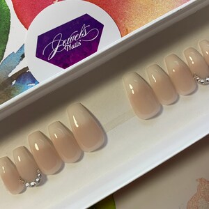 May include: Set of ten nude ombre coffin-shaped press-on nails with a single row of rhinestones on one nail. The nails are displayed in a white box with the text "Jewels Nails" on a purple sticker.