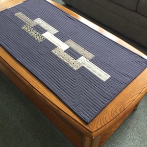 May include: A navy blue quilted table runner with a geometric design. The runner features a central pattern of rectangular shapes in shades of gray, white, and patterned fabric, set against a background of vertical stitching. The runner is placed on a wooden coffee table.
