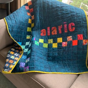 Pode incluir: Um edredom acolchoado para bebé em azul-petróleo com o nome "alaric" em letras coloridas. O edredom apresenta um padrão xadrez com quadrados de várias cores e uma borda com desenhos de elefantes. O edredom é adornado com amarelo.