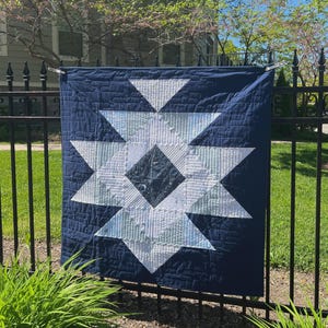 Könnte beinhalten: Eine marineblaue Steppdecke mit einem großen weißen Sternmuster. Der Stern besteht aus verschiedenen geometrischen Formen und Mustern. Die Steppdecke hängt im Freien an einem schwarzen Metallzaun, mit grünem Gras und Bäumen im Hintergrund.