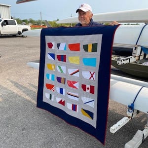 May include: A handmade quilt featuring colorful flag designs on a gray background, framed by a navy blue border with a red edge. The quilt is held up outdoors, showcasing its vibrant patchwork and nautical theme.