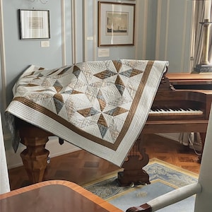 May include: A handmade quilt with a star pattern in shades of white, brown, and blue is draped over a wooden piano. The quilt has a brown border and is placed in a room with light blue striped wallpaper and framed artwork.