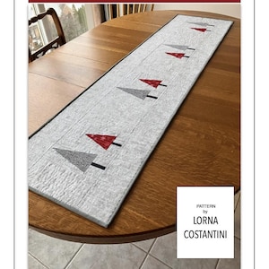 Può includere: Runner da tavolo rettangolare con sfondo grigio chiaro e motivo ripetuto di alberi di Natale stilizzati in rosso, grigio e nero. Il testo "Whispering Pines" è in alto. Le dimensioni finali sono 40,6 cm x 121,9 cm e 40,6 cm x 162,5 cm.