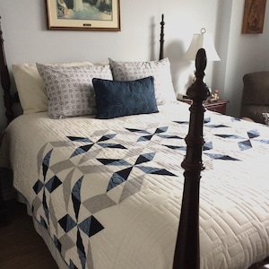 Puede incluir: Una cama con un edredón blanco con un patrón geométrico en tonos azules y grises. La cama tiene un marco de madera oscura y está adornada con almohadas en crema, gris y azul marino. Una imagen enmarcada cuelga en la pared sobre la cama.