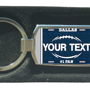 Könnte beinhalten: Ein silberner Schlüsselanhänger mit einem blauen und weißen Design im Fußball-Stil. Der Schlüsselanhänger trägt den Text "Dallas" und "#1 Fan". Der Text "YOUR TEXT" wird in einem weißen Oval auf dem Schlüsselanhänger angezeigt.
