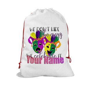 May include: White drawstring bag with a colorful Mardi Gras design. The bag features two theatrical masks in green, purple, and yellow, with the text "WE DON'T HIDE the crazy WE CELEBRATE IT Your Name". Red drawstring closure.