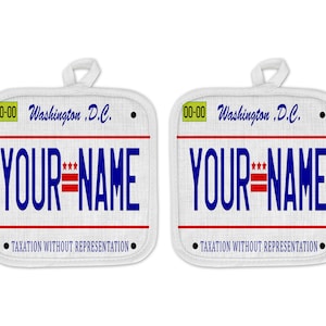 May include: A pair of white pot holders with a design that resembles a Washington, D.C. license plate. The license plate says "00-00 Washington, D.C." and "YOUR NAME" with a red, white, and blue American flag design. The bottom of the license plate says "TAXATION WITHOUT REPRESENTATION".