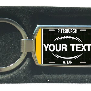 Könnte beinhalten: Ein silberner Metall-Schlüsselanhänger mit einem rechteckigen Anhänger mit einem Footballfeld-Design. Der Anhänger hat einen schwarzen Hintergrund mit dem Text "YOUR TEXT" und "#1 FAN". Oben auf dem Anhänger steht "PITTSBURGH".