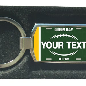Könnte beinhalten: Ein rechteckiger Metall-Schlüsselanhänger mit einem Footballfeld-Design. Das Design zeigt ein grünes Feld mit einem weißen Football und dem Text "YOUR TEXT" und "#1 FAN". Auf der Oberseite des Schlüsselanhängers steht "GREEN BAY". Der Schlüsselanhänger befindet sich in einer schwarzen Box.
