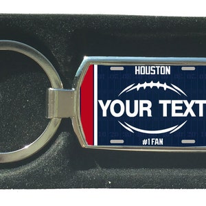 Könnte beinhalten: Ein silberner Schlüsselanhänger mit einem blauen und roten Nummernschild-Design. Das Schild zeigt "Houston" und "#1 Fan". Das Schild hat eine weiße Fußballgrafik und den Text "YOUR TEXT".
