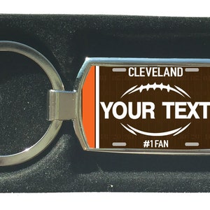 Könnte beinhalten: Ein silberner Metall-Schlüsselanhänger mit einem rechteckigen Anhänger, der ein braun-oranges Football-Design zeigt. Der Text "YOUR TEXT" ist prominent dargestellt, mit "CLEVELAND" und "#1 FAN". Der Schlüsselanhänger wird in einer schwarzen Box präsentiert.