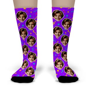 May include: A pair of purple socks featuring a repeating pattern of a smiling face, stars, and colorful confetti. The socks have black heels and toes. The socks are knee-high.