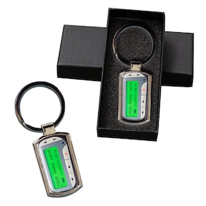 May include: Silver keychain with a digital display that says "YOUR NAME HERE 12:34PM 01/05/08". The keychain is in a black box with a black foam interior.