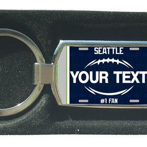 May include: A silver metal keychain with a blue and white football design. The keychain features the text "Seattle" and "#1 Fan". The text "YOUR TEXT" is also included on the keychain.