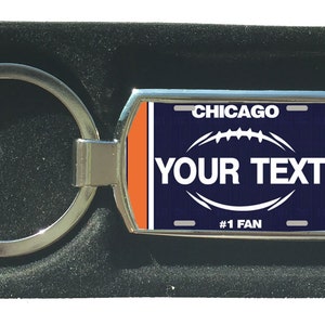 Könnte beinhalten: Ein silberner Schlüsselanhänger mit einem rechteckigen Anhänger in Blau und Orange. Der Anhänger trägt den Text "CHICAGO" und "YOUR TEXT" mit einer Fussballgrafik und dem Text "#1 FAN".