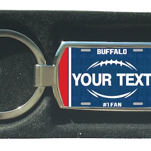 Könnte beinhalten: Ein silberner Schlüsselanhänger mit einem blauen und roten Design zum Thema Fußball. Der Schlüsselanhänger trägt den Text "BUFFALO" und "YOUR TEXT" mit den Worten "#1 FAN" unten.