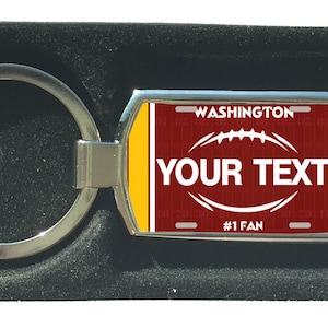 Könnte beinhalten: Ein silberner Schlüsselanhänger mit einem roten, weißen und goldenen Design einer Fußballl-Kennzeichenplatte. Die Kennzeichenplatte zeigt "WASHINGTON" und "#1 FAN". Der Text "YOUR TEXT" befindet sich in der Mitte der Platte.