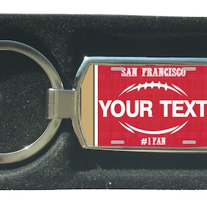 Könnte beinhalten: Ein silberner Schlüsselanhänger mit einem rot-weiß-goldenen Anhänger zum Thema American Football. Der Anhänger hat den Text "SAN FRANCISCO" und "YOUR TEXT" mit einer Football-Grafik und dem Text "#1 FAN".