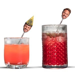 May include: Two glasses of red and orange cocktails with a person's face on a stick in each glass. The sticks have a party hat on top. The hats say "Happy New Year 2025".