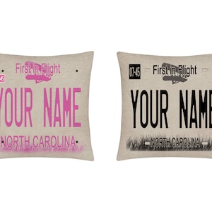 May include: Two decorative throw pillows with a vintage North Carolina license plate design. The pillows feature the text "First in Flight" and "Your Name" in pink and black lettering on a beige background.