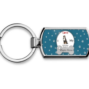 May include: A teal blue metal keychain with a silver ring. The keychain features a white bone with the text "I Love My Australian Shepherd" and a space for a dog's name. The background is covered in white paw prints.