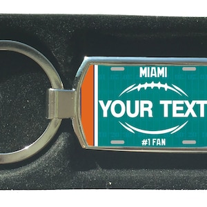 Könnte beinhalten: Ein silberner Schlüsselanhänger mit einem Fußball-Design in den Farben Türkis und Orange. Der Schlüsselanhänger trägt den Text "MIAMI" und "YOUR TEXT" sowie den Schriftzug "#1 FAN" am unteren Rand.