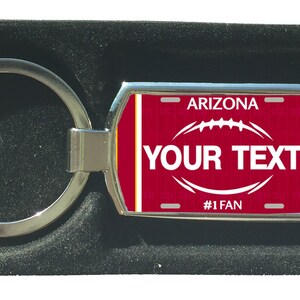 Könnte beinhalten: Ein silberner Schlüsselanhänger mit einem roten und weißen Arizona-Kennzeichen-Design. Das Kennzeichen trägt den Text "Arizona" und "#1 Fan" sowie eine Fußballgrafik. Der Text "YOUR TEXT" befindet sich in der Mitte des Kennzeichens.